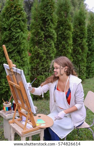Female artist holds brush and wrote oil on canvas painting, does what likes, interests and hobbies,  preparing picture to order, advertises or art studio for drawing utensils, sits on chair at  wood