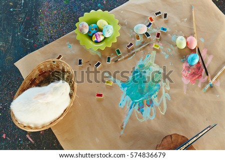 White rabbit in basket, paints, paintbrushes and Easter eggs on sheet of paper