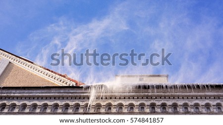 wind blows to roof of old house and snow fly to ground against blue sunny sky with white clouds. Heavy snowfall on a city house roof in the winter time of the year
