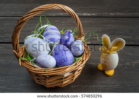 Easter wicker basket with colored eggs and a small egg rabbit on grey wooden board.