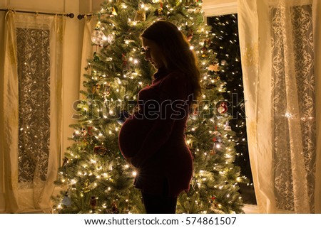 A pregnant women stands in front of a christmas tree with lights on in low light making a beautiful silhouette