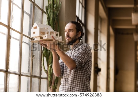 Young architect is looking on the new model in the old industrial space with big factory windows. Man is standing in front of window and holding small model of the house. Color toned image. Royalty-Free Stock Photo #574926388