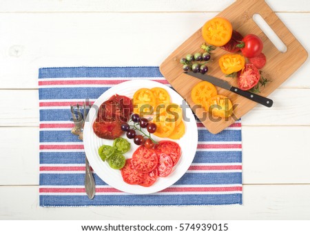Bright and Colorful Organic Tomato Variety sliced on a plate and red, white, and blue striped placemat centered on Rustic Wood Board Table Background with cutting board and knife