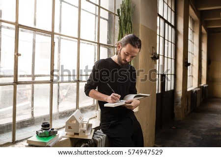Young architect is writing or drawing on his notebook in the old industrial space with big factory windows. Man is sitting in front of window and holding the sketch book. Color toned image.