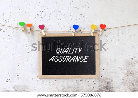 Quality Assurance - concept words on blackboard with wooden clamps on rustic wooden background.