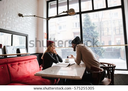 Side view of woman sitting by the table with man in cafe and man showing photo on smartphone