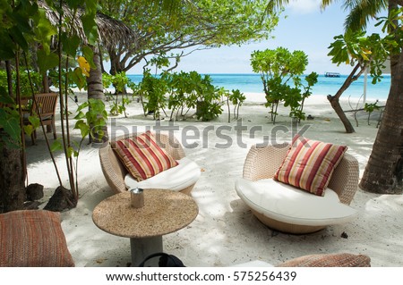 Beach paradise island in Maldives. Two chairs with cushions with luxurious drapes and table for guests in the foreground. In the distance the turquoise sea and green palm trees , white sand. . Royalty-Free Stock Photo #575256439