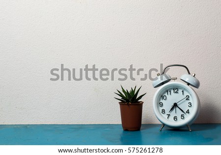Workplace with table clock, plants on blue table. White wall.
