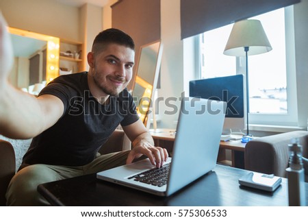 Young man sitting at a laptop a freelancer in the office does selfie. Home Office. Royalty-Free Stock Photo #575306533