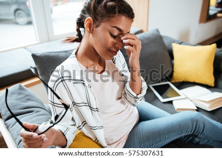 Photo of young tired african lady holding glasses sitting in library touching nose.