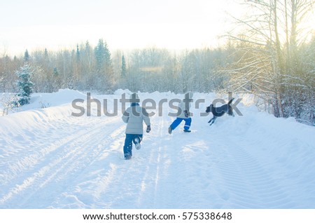 two boys dressed in grey jackets and blue pants and black dog run and play on rustic snow-covered country road at Sunny day on background of a winter forest.
