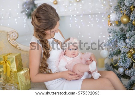 Cute mother mom in fairy airy dress with beautiful daughter celebrating Christmas together close to xmas tree with presents.Brunette curly hair cozy dresses woman holding baby in her hands.