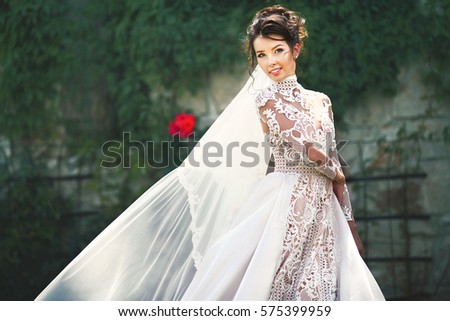 Wind blows bride's veil while she poses before the wall covered with green ivy and red flowers