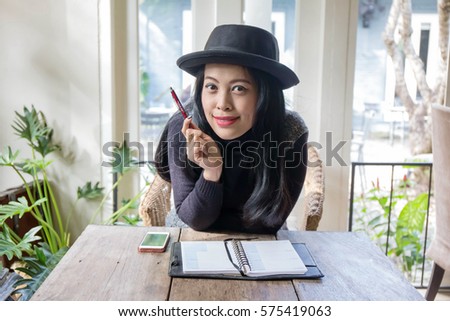 Charming beautiful tan skin Asian business chic woman hand work on laptop phone and write pen on notebook dairy on wooden table in coffee shop . Presenting your product with good looking woman face.