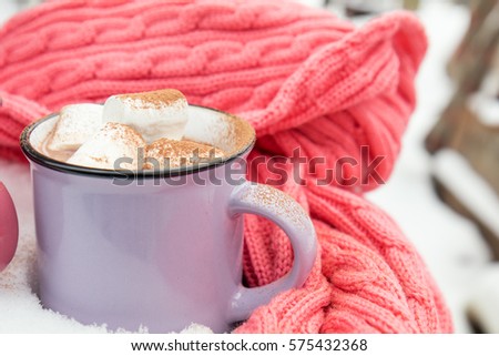 Hot chocolate with marshmallow in violet cup wrapped in a cozy winter pink scarf on the snow-covered table in the garden. Coloring and processing photo, selective focus, small depth of field.
