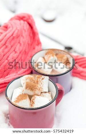 Hot chocolate with marshmallow in pink and violet two cups wrapped in a cozy winter pink scarf on the snow-covered table in the garden. Coloring and processing photo, small depth of field.