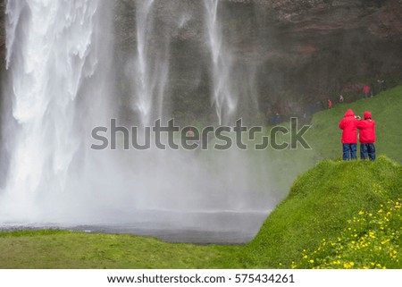 Two tourists taking pictures at Seljalandsfoss waterfall in Iceland dressed in red rain coats with mist in distance