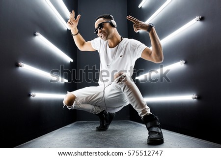 young handsome hipster man listening to music on headphones, smiling happy, white apparel, disco night club, having fun, hip hop style, positive mood, face expression, dancing Royalty-Free Stock Photo #575512747