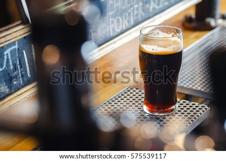 Nonic pint glass with dark stout ale standing on a pub counter near draft taps Royalty-Free Stock Photo #575539117