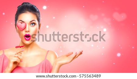 Beauty surprised Young fashion model Girl with Valentine Heart shaped cookie in hand. Love. Beautiful young woman pointing hand, advertising gesture. Valentines Day gift. Pink polka dots background