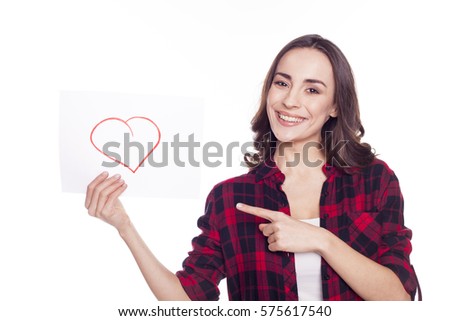 Young beautiful girl in a red checked shirt smiles and holds in her hands a white paper with with a draw heart on a white background isolated. With love. The 14th of February.