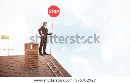 Engineer man standing on house roof and holding red prohibition sign. Mixed media