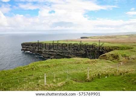 Cliffs covered in green grass at the Irish coast on a blue and cloudy afternoon in a panoramic landscape picture with a fence in the foreground and the sea in the background.