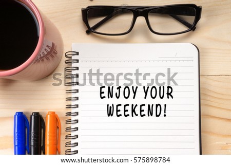 ENJOY YOUR WEEKEND! on notebook with cup of coffee and glasses on wooden background.