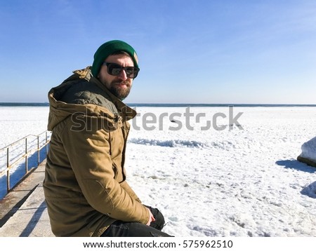 Cheerful bearded man looking to a camera.man in a green hat and jacket sitting on a pier.Stylish winter wear.black jeans,man in sunglasses,man on the sea,stylish outfit,attractive,casual wear