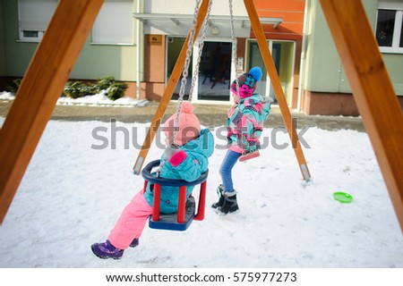 Two little girls swinging on a swing at the playground in the courtyard of an apartment building. Ground is covered with snow. Children dressed in warm ski suits. A fine winter day.