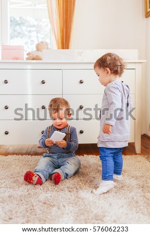 Happy little boy sitting on the floor and playing with smartphone and little girl standing behind and looking at him