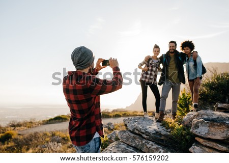 Young man taking picture of his friends while trekking in countryside during summer vacation. Group of people on hiking taking photographs with mobile phone.