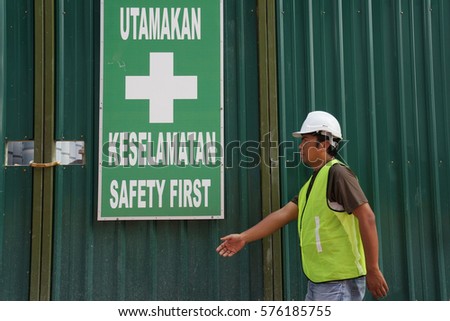 Worker wearing safety helmet and jacket at working area with the sign board of  'SAFETY FIRST'. Words 'UTAMAKAN KESELAMATAN' is malay language which mean 'SAFETY FIRST' in English