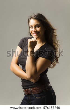Young mixed race woman wearing a black t shirt and jeans stands with hair blowing and hand on chin while smiling at viewer