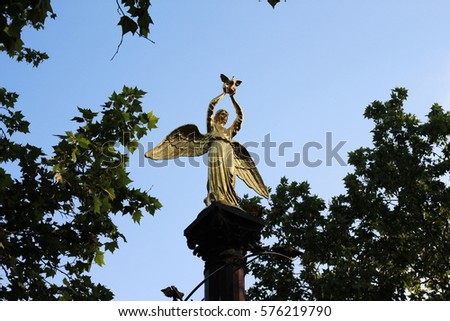 Photo golden statue of an angel with a dove in his hands on a pedestal in the form of a column on a background of blue sky and foliage of trees.