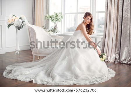 Wedding. Bride in beautiful dress sitting on sofa indoors in white studio interior like at home. Trendy wedding style shot in full length. Young attractive caucasian brunette model like a bride Royalty-Free Stock Photo #576232906