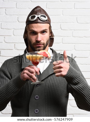 handsome bearded aviator man with long beard on serious face holding glass of alcoholic cocktail in gray knitted sweater with hat and glasses on white brick wall background