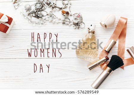 happy women's day text sign. luxury expensive jewelry and make up essentials and perfume flat lay on white rustic wooden table with space for text in soft morning light.  modern woman set