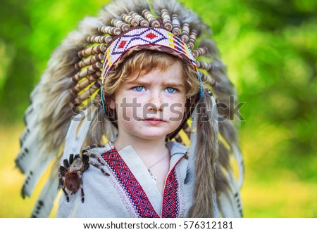 boy and spider on his shoulder, arachnid, blond boy in traditional American headdress, child and tarantula, the boy's face with freckles, headdress with eagle feathers, greenery background.