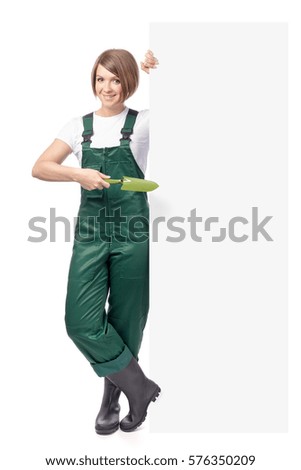 young smiling woman professional gardener with trowel standing next to the banner with empty copy space isolated on white background. advertisement blank board. gardening service and business concept