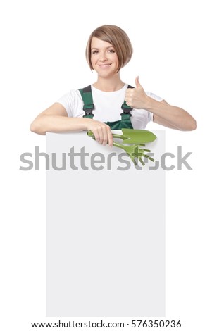 young smiling woman professional gardener with gardening tools showing thumb up and peeking from behind the banner with empty copy space isolated on white background. advertisement blank board