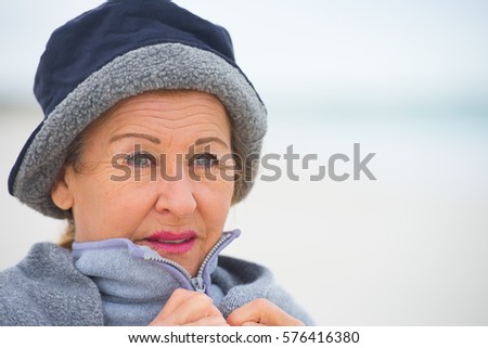 Portrait attractive senior woman at ocean, feeling cold and frosty, wearing jumper and hat to keep warm outside, copy space and blurred background.