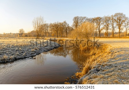 Picturesque rural winter landscape with a thin layer of snow and hoarfrost and a bent stream in low early morning sunlight.