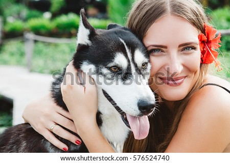 Young blonde woman in black dress with red hibiscus flower with husky dog at sunny weather, friendship between human and animal