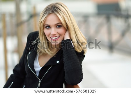 Attractive blonde woman smiling in urban background. Young girl wearing black zipper jacket sitting on a bench in the street. Pretty female with straight hair hairstyle and blue eyes.
