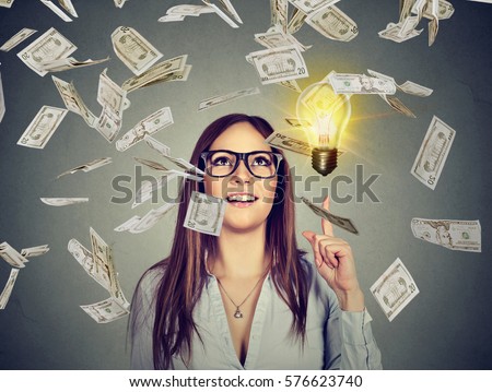 Portrait happy woman in glasses has a successful idea under money rain isolated on gray wall background with bright light bulb above head 