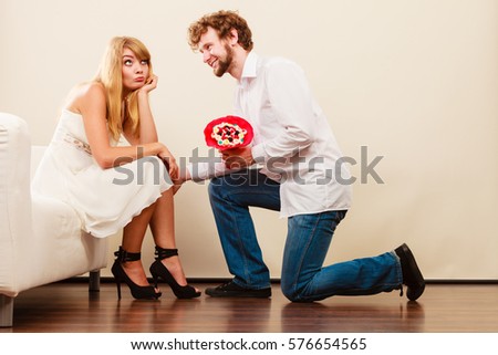 Handsome man giving pretty unhappy bored woman candy bunch flowers. Young boyfriend with present gift kneeling in front of girlfriend.