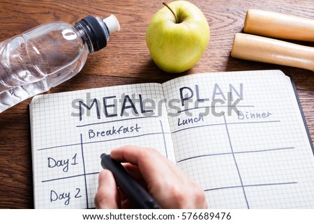 High Angle View Of A Person Hand Filling Meal Plan In Notebook At Wooden Desk Royalty-Free Stock Photo #576689476
