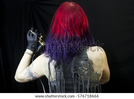 Tattooed girl with red and violet hair in different poses with tattoo machine as tattoo master. Master works. Black background and sterile black gloves. Emotional face. Tattoo glasses.
