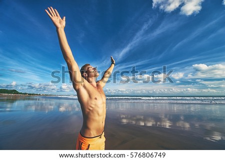 Victory and freedom. Young handsome strong man raising hands up on the beach against sky.